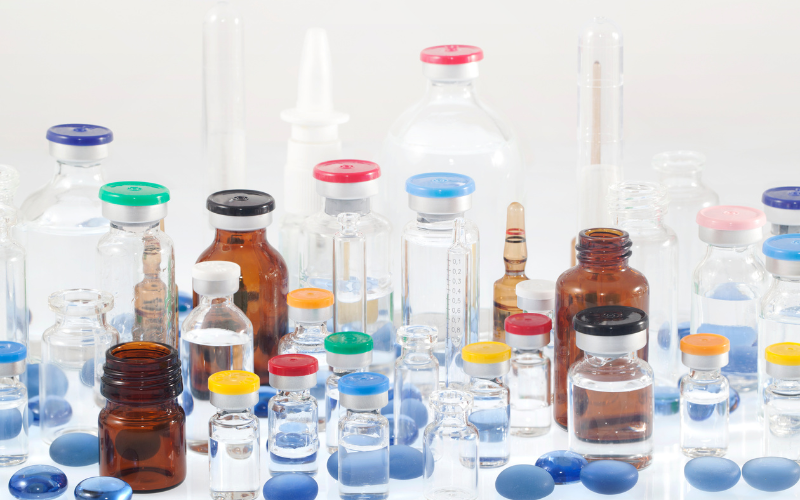Assorted medical vials, ampoules, and pill capsules arranged on a white surface, with colorful caps and various liquid contents.