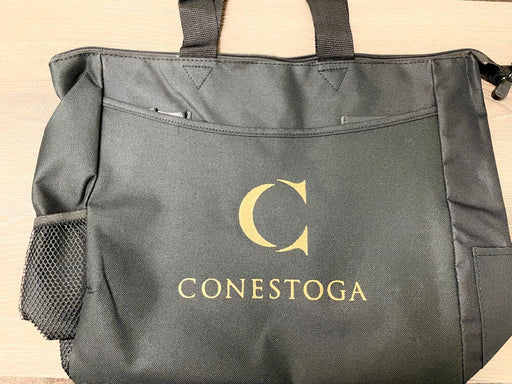 A black tote bag sits on a surface. The bag features a gold "C" and the word "CONESTOGA" embroidered on the front.